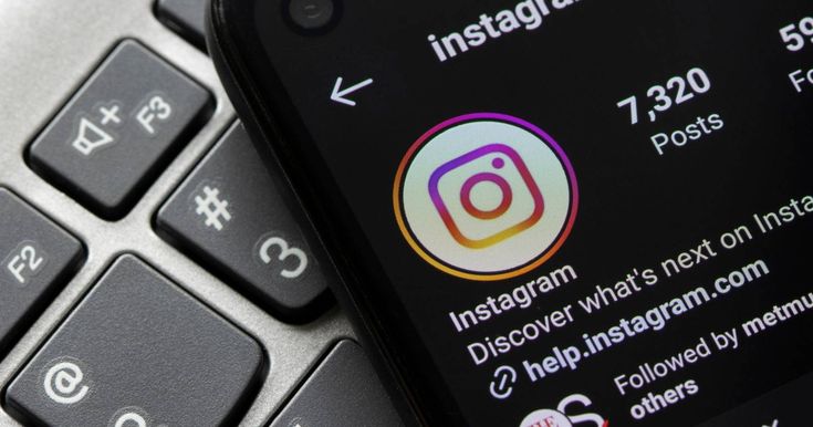 A close-up shot of a smartphone lying on a grey computer keyboard. The phone is in dark mode and displays the official @instagram profile page. The colorful Instagram logo is visible as the profile picture, along with the stats showing 7,320 posts and a link to help.instagram.com in the bio.