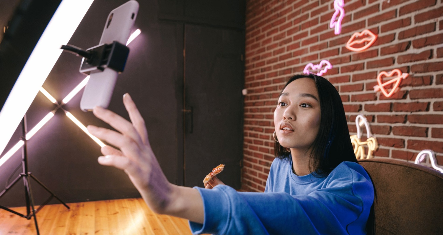 A young woman in a blue sweater holding a slice of pizza while reaching toward a smartphone mounted on a ring light to record an Instagram Reel in a room with a brick wall and neon signs.