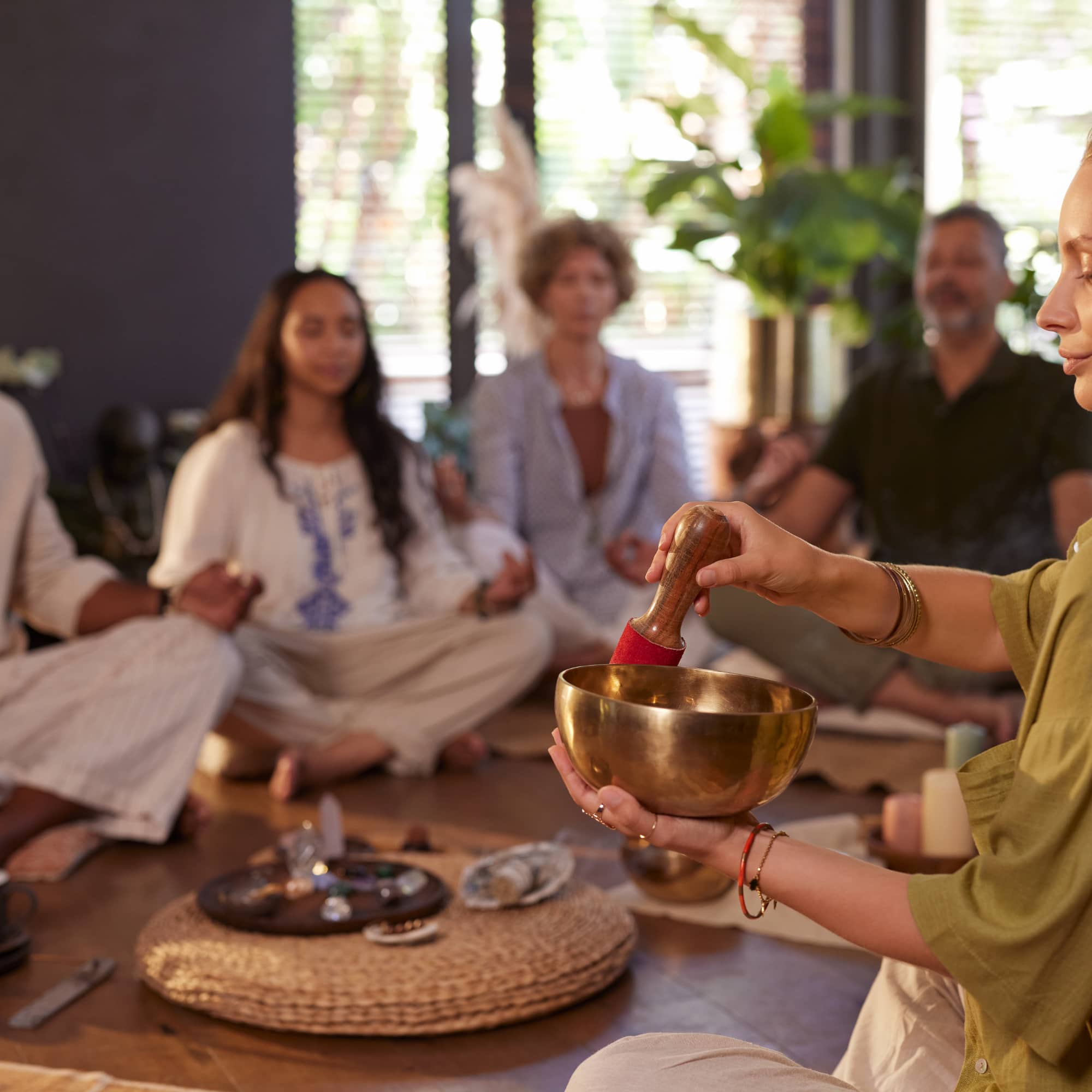 A person plays a singing bowl during a group meditation session, with participants sitting cross-legged in a calm indoor setting.