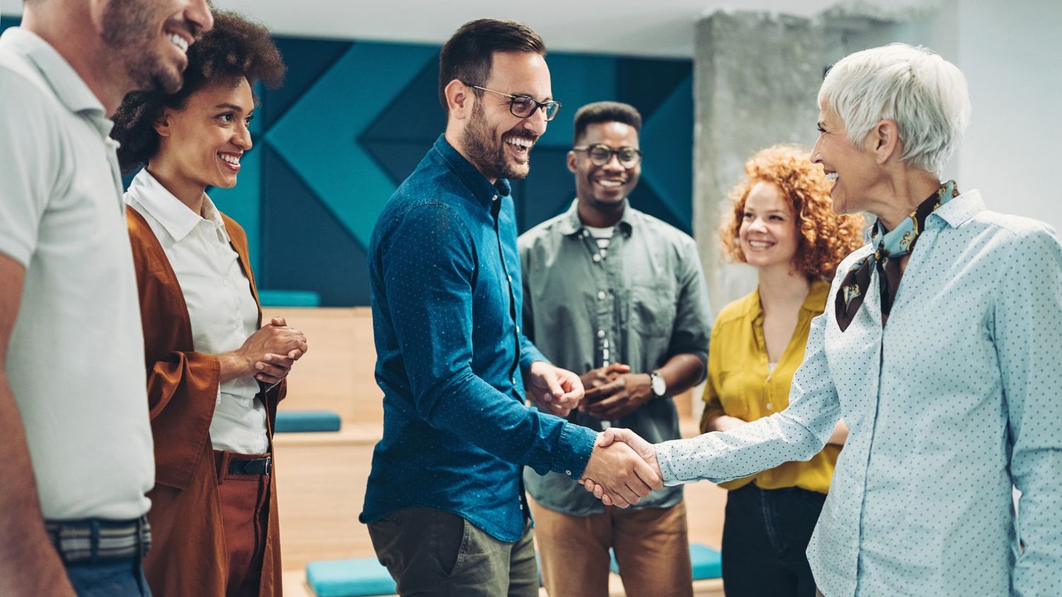 A diverse group of smiling professionals standing in a modern office, with a man and woman shaking hands in the foreground.   The image captures a warm welcome and positive introduction, representing the importance of a strong first impression through a welcome message.