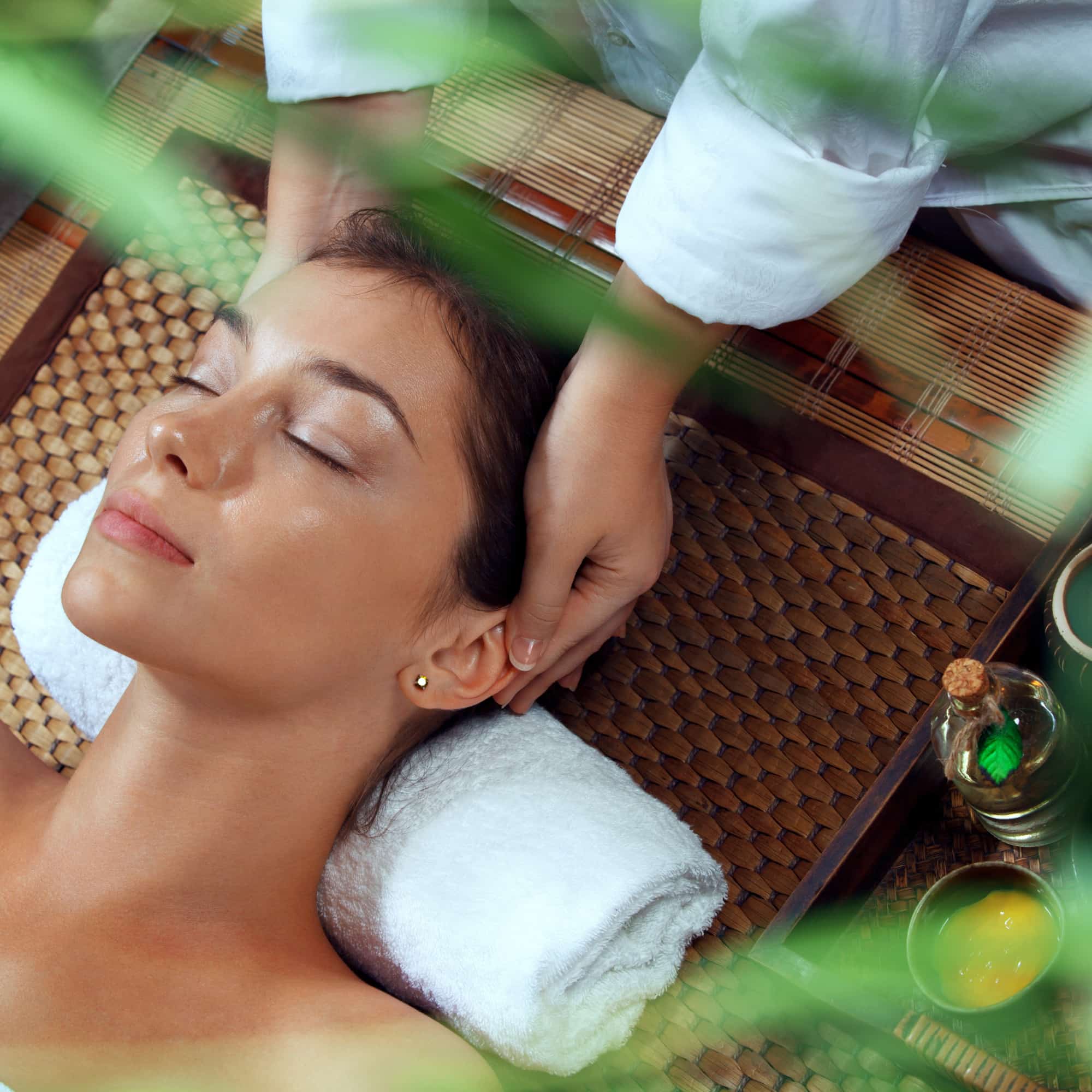 A woman relaxing with eyes closed while receiving a head and neck massage at a spa, surrounded by natural elements and skincare products.