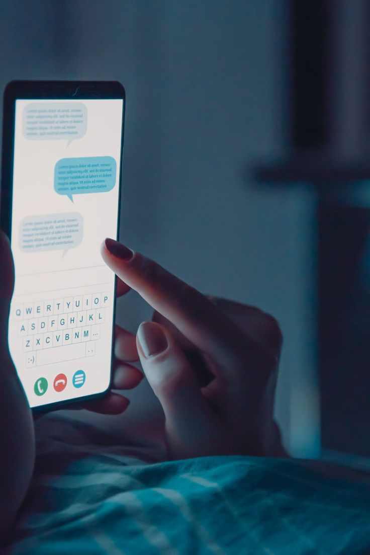 A close-up of a person's hands holding and interacting with a brightly lit smartphone in a dimly lit room, likely lying in bed. The phone screen displays a generic messaging application interface showing chat bubbles and a QWERTY keyboard. The person's index finger is touching the screen.