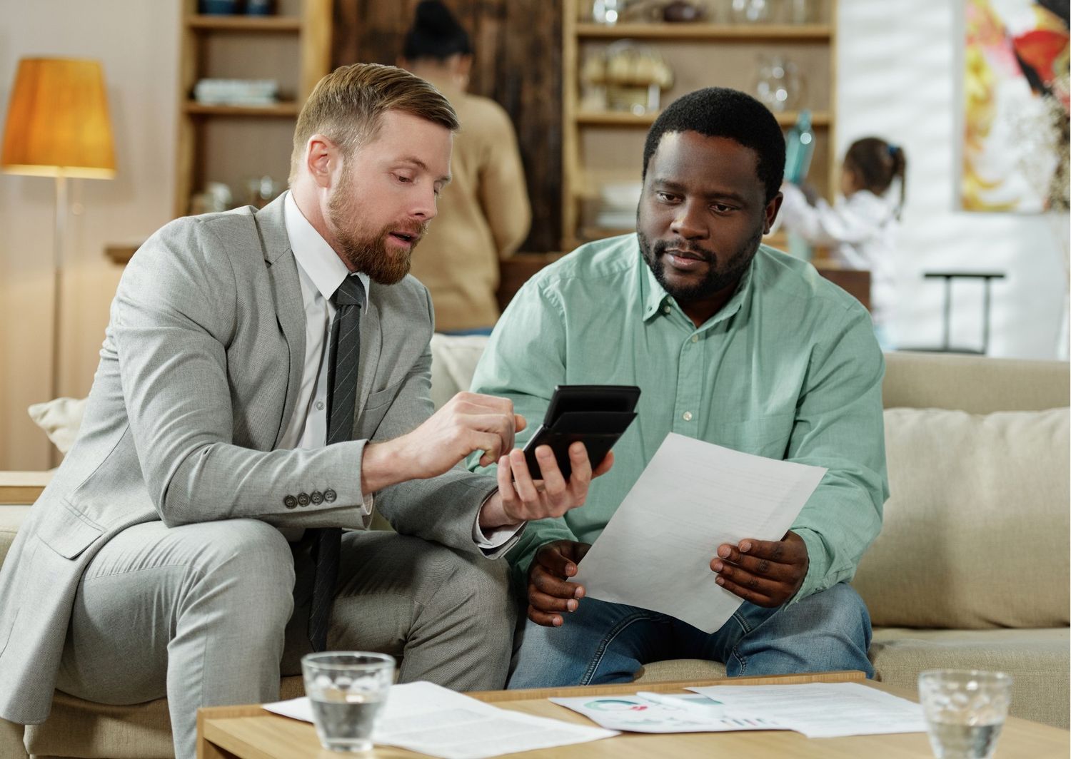 This image shows two men sitting on a couch, deeply engaged in a discussion over financial documents and a calculator. The setting is a warm, home-like office environment, which adds a sense of personal trust and consultation to the professional interaction.  In the context of the digital marketing and SEO series you've been building, this visual represents the human element of the "close"—the point where data meets personal decision-making.