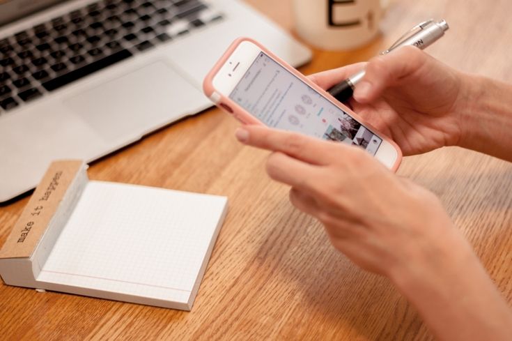 A person’s hands hold a pink-cased smartphone and a silver pen over a wooden desk equipped with a laptop and a 